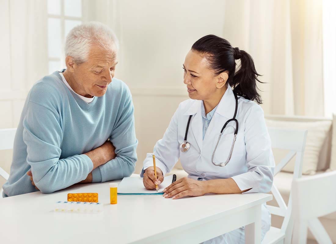 Medicare Part D - Smiling and Encouraging Doctor Takes Notes as She Discusses Medication Precautions and Side Effects with a Senior Male Patient in His Home at the Kitchen Table